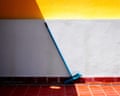 A broom leaning at an angle against a wall, its stick aligning with the shadow on a yellow wall above, with red tiles below