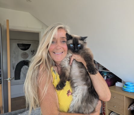 A woman with a yellow vest top smiles as she hugs a very fluffy brown and grey cat with blue eyes.