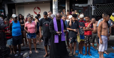 A priest stands over a line of bodies as onlookers listen to his address.