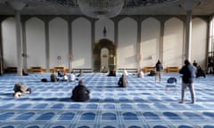 About a dozen people sit, pray and stand in a blue carpeted hall with arched walls and a large light fitting in the centre