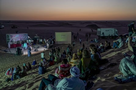 People seated on the sand at dusk watch an open-air concert performance