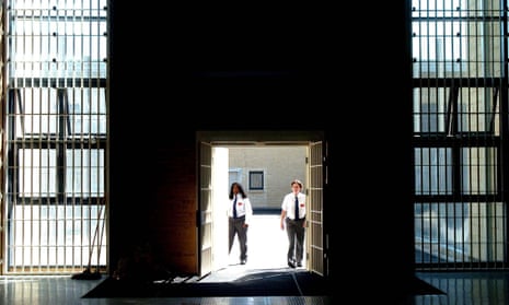 View out from inside a British prison – HMP Bronzefield, a women's prison in Surrey – through open gates with sunlight streaming through the aperture and the barred windows to either side. Two female prison guards stand in the gateway.