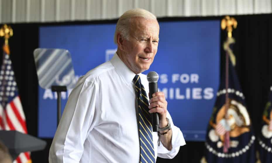 Joe Biden, in a dress shirt with the sleeves rolled up, speaks into a microphone in front of an American flag and a flag bearing the presidential seal.