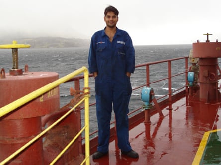 Bahri stands on the deck of a ship wearing a blue boilersuit, hands in pockets.