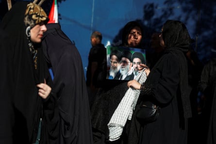 Women wearing head coverings stand together as mourners gather in Mashhad to mark the burial of Ali Khamenei. One woman holds a photograph showing the late Ayatollah Ruhollah Khomeini, Khamenei, and Iran’s new supreme leader, Mojtaba Khamenei.