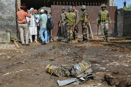 Five soldiers stand next to a small group of civilians by wall with exploded shell in foreground