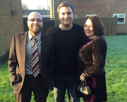 Dylan and his parents stand on grass near their house. He is taller than both of them and has a cheerful face and short light brown hair. Karen has dark, shoulder-length hair at this time and Tosh a beard. They look dressed up as if to go out, with Karen in a brown velvety dress and Tosh in a suit and striped tie. All three look happy and smiling.