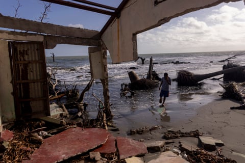 A view from inside the remains of a building, with the roof and much of the walls missing, of uprooted trees and a boy running out of the waves on the shoreline