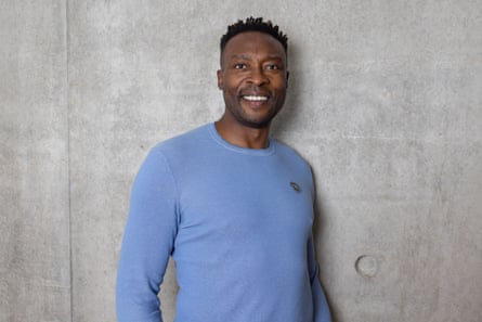 Former Newcastle striker Shola Ameobi smiles, wearing a light blue jumper against a concrete wall