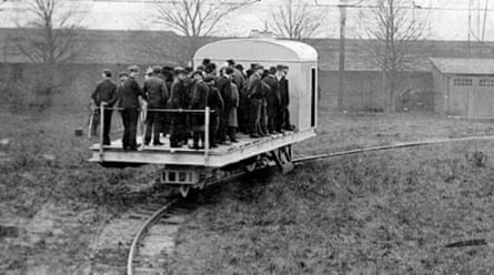 Men stand on the platform at the rear of a monorail that is moving through a field. It looks broadly like a large milk float that hasn’t a roof