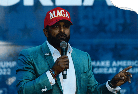 a man in a suit and a red Maga cap speaks into a microphone