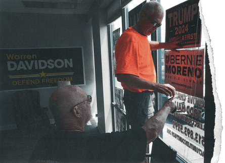 Two men hang campaign signs in a window, one reading ‘Bernie Moreno for US Senate’ and the other ‘Trump 2024 America First’.