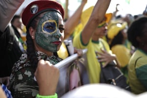 A close-up shot of a woman in a red beret with half of her face painted with the Brazilian flag with other protesters in the background