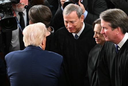 Two men, one with a unique hairstyle and the other in a judge’s robe, shake hands and speak with each other while other people in judge’s robes gaze obsequiously at the person with the floofy hairdo.