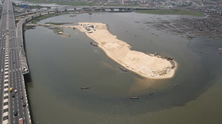 ‘The water is no longer our friend’: how dredging is pushing Lagos Lagoon towards ecosystem collapse – photo essay A large sandbank rises out of body of water. A four-lane highway runs along the lefthand side looping round to the right in a slip road. On the right are some floating houses.