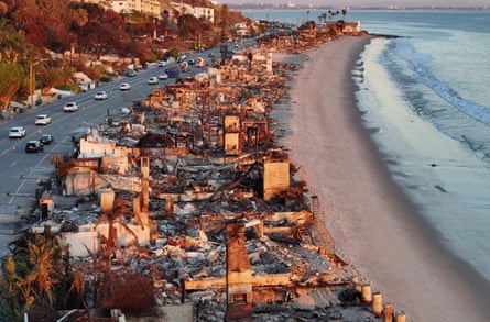 Aerial view of burnt down beachfront homes in ruins; vehicles travel on a road to one side and there is a long sandy beach and sea on the other.