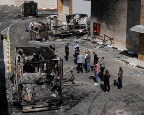 A group of about a dozen people stand outside near the charred remains of trucks