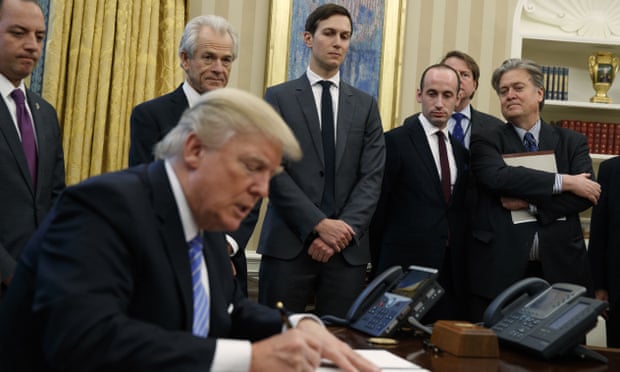 Donald Trump sits at a desk in the Oval Office signing a document. Behind him are members of his cabinet.