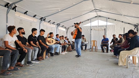 A teacher with a guitar stands between rows of seated music students in a tent