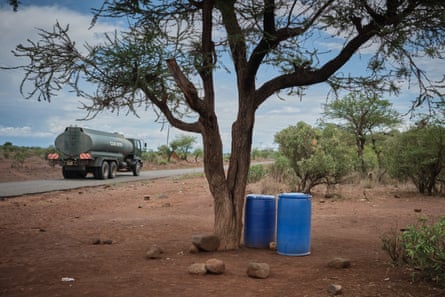 A truck with “clean water” written on the side passes by water drums under a tree at the roadside