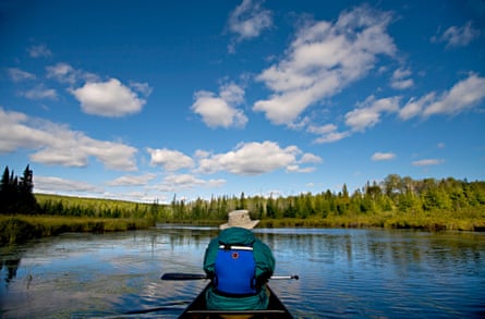 Taken from back of kayak, person in hat and jacket with oar on lap floats on blue lake under partly cloudy sky.