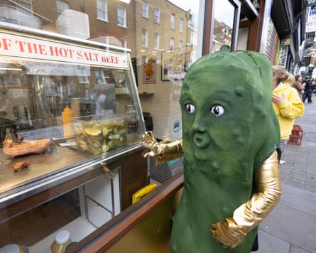 Person in green pickle suit beside a hot salt beef counter with real pickles on urban street in London