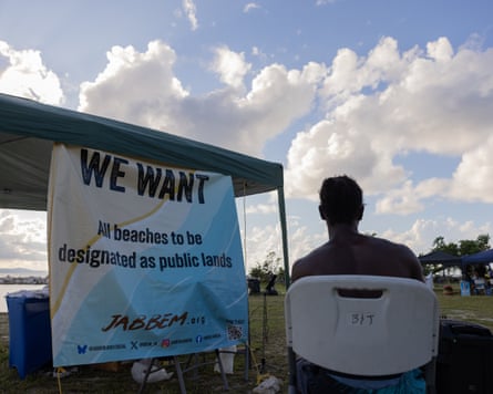 A man sits before a banner reading ‘We Want All beaches to be designated as public lands’ in Montego Bay