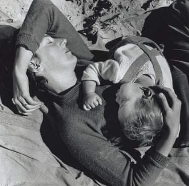 A mother lies on sand with eyes closed, cradling her baby against her chest