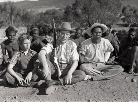 A white man with a rifle and a child beside him sits in front of a group of Indigenous people