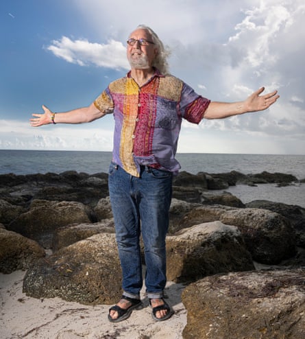 Billy Connolly standing on a rock, sea and blue sky behind him, arms reaching out to the side