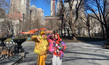 two people in brightly colored jackets, one holding a dog, do a thumbs down in front of the skyline
