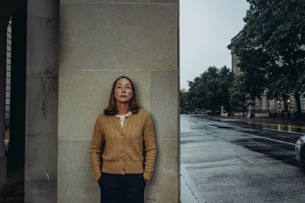 Kathleen Walters wearing a beige cardigan with a white top underneath and dark trousers, standing with her hands in her pockets against a concrete wall on the side of a road