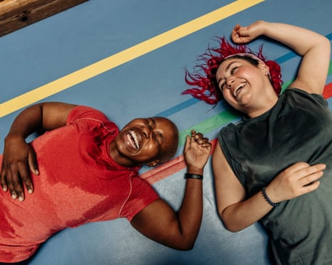 Two female athletes lie laughing on a sports court safety mat, arms raised