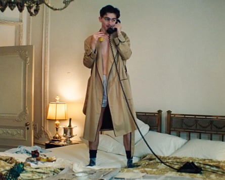 A man wearing nothing but glasses, boxers and a trenchcoat stands on a bed making a phone call