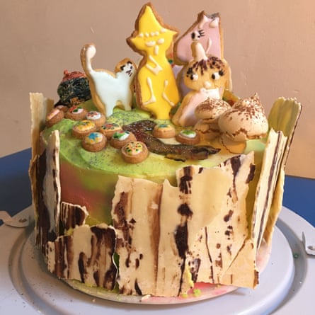A mish-mash of yellow white and pink iced biscuits shaped like Moonmintroll characters and mini cupcakes adorn the top of a tall iced rainbow coloured cake