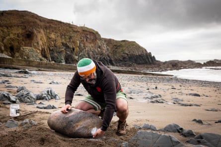 Keohan brushes sand away from a stone with his hands on a beach, before lifting it