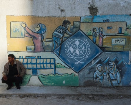 A man sits next to a wall on which a mural has been painted depicting United Nations Relief and Works Agency staff at work