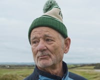 Bill Murray in a green bobble hat and blue jacket looks off to the side outdoors