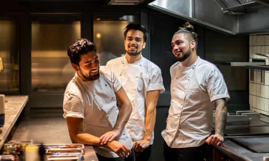 The three brothers smiling in their chef’s whites as they lean on tables and cookers in their kitchen