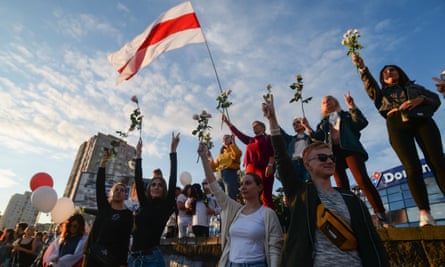 A group of protesters holding flowers in their raised hands with the flag of Belarus in the background