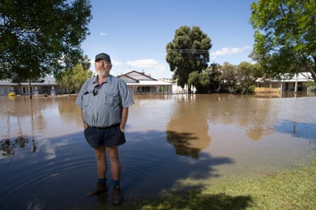 A man stands at the edge of brown flood waters that have covered a residential street, with houses in the background