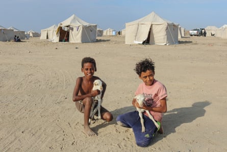 Two children sit with small goats with tents in the background