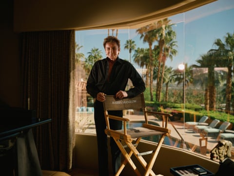 Barry Manilow standing behind director’s chair with large windows behind him and a view of palm trees