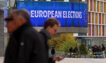 A sign reading 'European Elections' is displayed on a building while people walk in front of it in the foreground