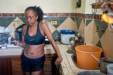 A woman in sports bra and shorts, holding a cigarette, while water flows from a tap into a large orange bucket in her kitchen sink.