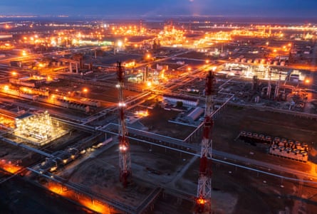 acres of illuminated pipes, towers and storage facilities stretch toward the horizon at dusk, the lights glowing orange and yellow like flames against the dark-blue sky.