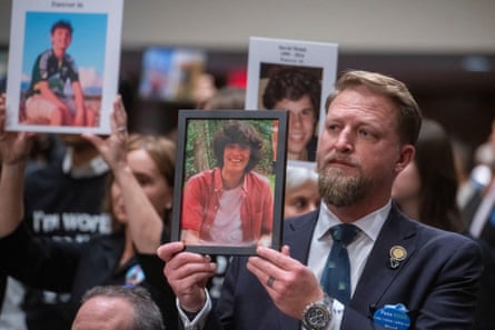 A man in a suit holds up a framed photograph of his son. People behind him are also holding up portraits of teenage boys