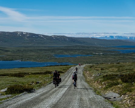 Cyclists ride along a gravel road towards lakes and mountains under a blue sky