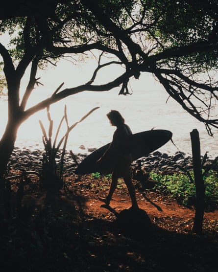 A person carries a surfboard along a dirt track with water in the background