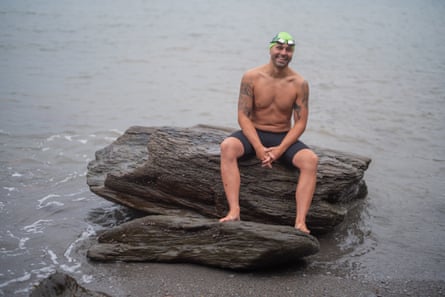 Neil Gilson sits smiling on a rock on a beach. The water and sand look dark and grey. He wears black shorts and a bright green rubber hat with goggles on his forehead.
